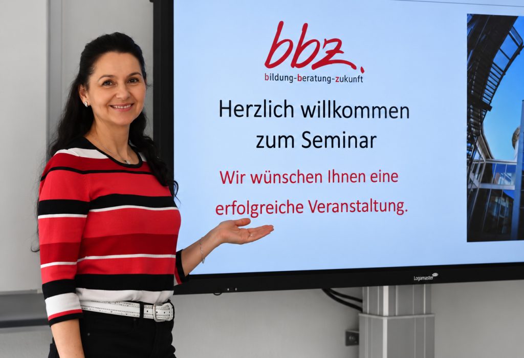 Frau steht vor Leinwand mit BBZ Logo und Willkommensgruß für Seminar